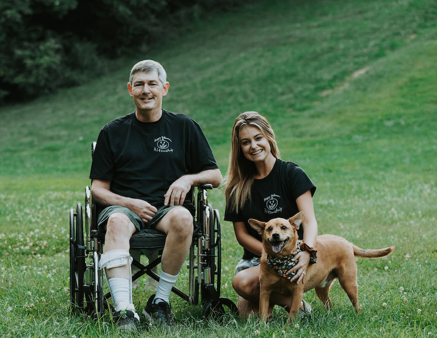 A photo of a man in a wheelchair beside a woman with her arms around a dog. They are both smiling and are seated on a grassy hill.