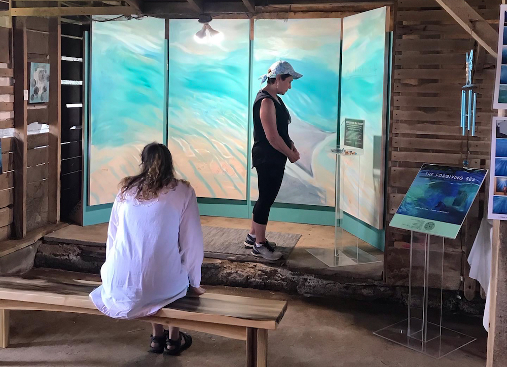 Two women contemplate a large scale painting of ocean waves viewed from in the water.
